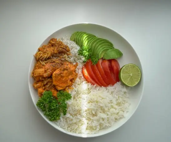 Foto de um prato de comida no refeitório da empresa, o prato está sobre um tampo branco e possui uma porção de arroz, estrogonof, salsa, tomate, abacate fatiado e uma fatia de limão