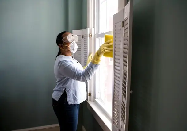 Funcionária de empresa de facilities, mulher de camisa branca de manga comprida em pé, com máscara de óculos e proteção limpando uma janela com a moldura de madeira branca