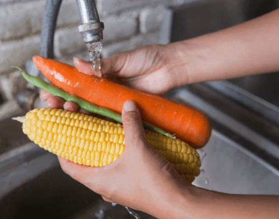 foto de par de mãos lavando legumes (cenoura, vagem e espiga de milho) em uma torneira aberta