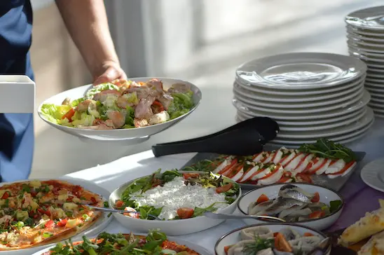 Foto do braço de uma pessoa próxima à um buffet do que seria um restaurante corporativo, se servindo de pratos feitos com saladas frias, tomates e mozzarella de búfala