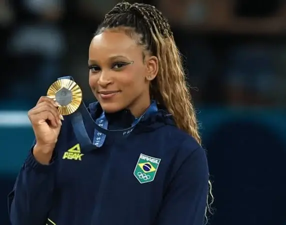 Foto da ginasta Rebeca Andrade sorrindo, segurando sua medalha de ouro conquistada nas Olimpíadas de Paris 2024 à altura do seu rosto