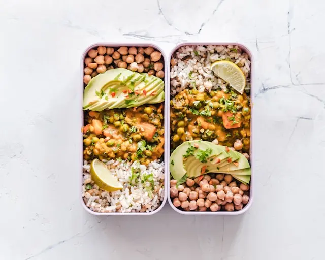 Foto de refeição transportada em duas marmitas, com arroz, grão de bico, limão, abacate e outros alimentos.