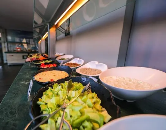 Foto tirada da ponta de uma mesa de um buffet de saladas de um restaurante interno da empresa. Na imagem, é posível visualizar as opções de saladas e molhos e a cozinha bem ao fundo da imagem na outra ponta da mesa