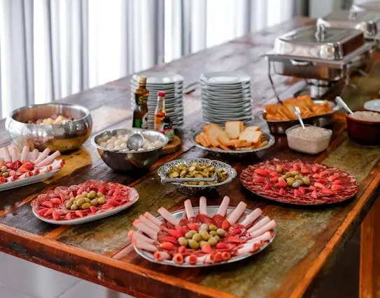 foto de um buffet de frios e panelas de inox para manter pratos quentes sobre uma mesa de madeira, os pratos de frios e quentes estão bem alinhados ao longo da mesa retangular