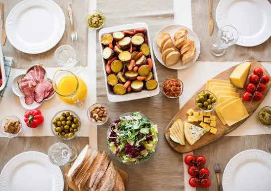 Foto de cima de uma mesa posta com 4 pratos brancos e talheres, com alimentos servidos no meio da mesa como tábua de queijos, salada de folhas, jarra de suco de laranja, pães fatiados e batata doce cozida, oferecendo uma alimentação terceirizada variada e completa.