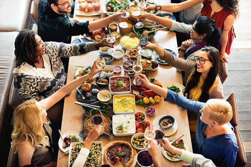 Pessoas reunidas em torno de uma mes de madeira, com comidas, todas brindando com seus copos e taças