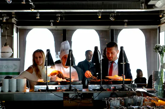 Imagem de um buffet com um homem de terno e uma mulher se servindo, entre eles está o chef de cozinha, há um garçom de pé ao fundo em um ambiente de alimentação corporativa