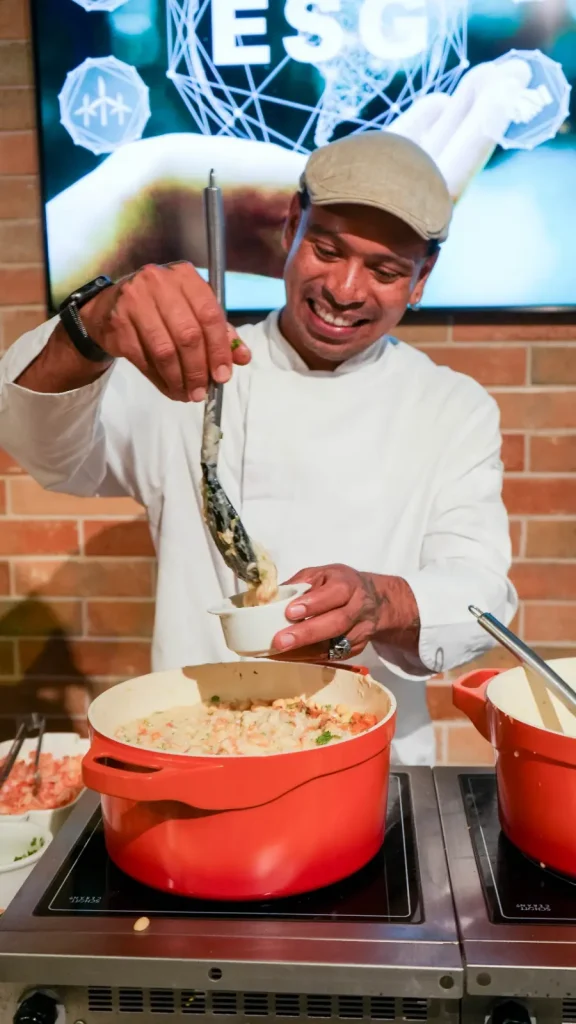 Foto do Chef Cadu de Oyá cozinhando. O homem está sorrindo e olhando para a comida. Se trata de um homem alto, afrobrasileiro, vestindo um boné e uma doula sorrindo e manipulando uma colher em uma panela sobre o fogão